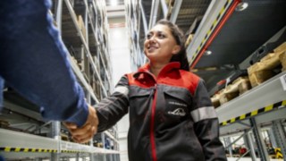Eine lächelnde Servicetechnikerin von Linde MH schüttelt die Hand einer anderen Person zwischen Lagerregalen.