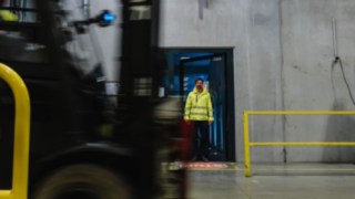Ein Mann in Sicherheitskleidung tritt aus einer Tür in eine Werkshalle. Ein Stapler fährt vorbei.